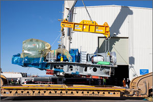 This photo shows the enormous blue and white cylindrical drive train of Samsung's wind turbine being unloaded by a crane from an extra-long semi-trailer outside the dynamometer test facility.