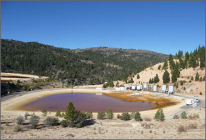A photo of a lake with purplish-yellow water with small mountains dotted with evergreens in the background.