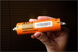 Photo of a lithium-ion battery meant for an automobile shows a hand holding an orange cylinder with A123 Systems and other writing on it. The battery's bolt-like terminals protrude from each end. 