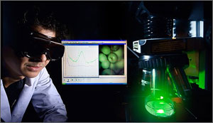 Photo of a man in a lab wearing goggles with computer monitor behind him and a microscope device with a green laser.