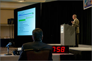 Photo of a man standing onstage and giving a presentation. In the foreground, a man sits next to a large digital clock. 