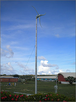Photo of a grayish wind turbine seemingly growing out of a flower garden and soaring above low-rise classroom buildings, with cumulus clouds in the background.