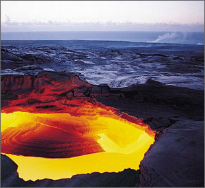 Photo shows fiery hues of red and yellow rising from a circular vent, with hardened lava in the background, giving way to the sea.