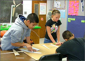 This photo shows a colorful classroom with a boy and girl using short-bladed knives to cut turbines out of balsa wood.