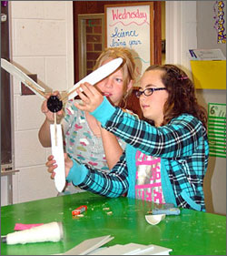 This photo shows two girls holding up their blade design and intently examining it. The turbine's blades are glued to wooden rods that are attached to a hub. The girls are standing at a bright green table.