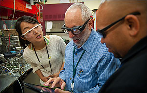 In a photo, three scientists talk as principal investigator David Ginley holds an I-pad version of a state-of-the-art periodic table
