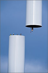 In this photo, the heads and torsos of four workers can be seen near the top of a white cylindrical tower piece. They're eying the next section that is being lifted by a crane.