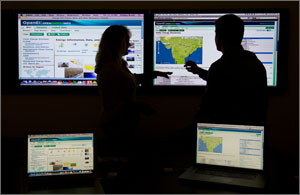 In a photo, a woman and man in silhouette point to brightly lit web pages projected on a large white screen in a small room. Directly between them is a screen shot of a map of India.