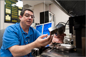 In this photo, a scientist in a blue polo shirt is sitting in front of a huge microscope, holding a probing device.
