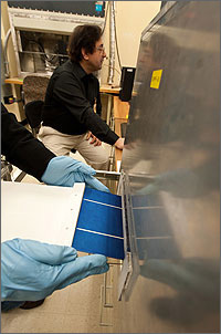 This photo, a close-up of hands, protected by sanitary blue plastic gloves, are installing a square solar wafer into the Optical Cavity Furnace, much like someone would install a CD into a CD player. In the background is Bhushan Sopori sitting at his computer screen.