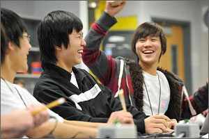 Photo of three young men while participating in science bowl competition.  One has arm raised in the air in celebration of correctly answering a question while the other two are laughing.