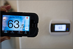 This photo is a close-up of a smart phone tuned to an application that can read and monitor a home's internal temperature from anywhere an Internet connection can be made. In the forefront the smart phone is held by hand and reads 63.5. In the background the thermostat on the wall also reads 63.5 degrees.