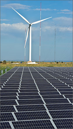 In this photo, rows of angled, bluish solar modules dominate the foreground, while a large, white wind turbine looms in the back under a blue, partly cloudy sky.