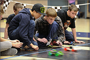 In  this photo, two middle-school boys are crouched down on a strip of neoprene  rubber watching their red car take off at the start of a car race. A green car  is next to it, neck and neck.