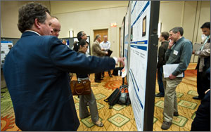 In this photo, people are perusing posters set on easels in a hotel ballroom setting. The posters contain information about how to improve the reliability of solar panels.