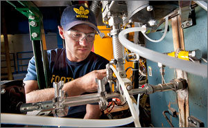 This photo shows a man in a blue T-shirt, baseball cap and safety glasses adjusting settings on a stainless steel instrument with a two-foot-long auger in the foreground.
