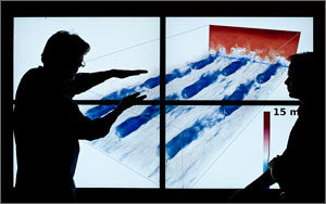 Two men in silhouette stand in front of a screen and demonstrate a computer simulation. On the screen is a computer simulation showing how the wind flows through a group of wind turbines.