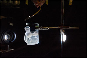 In this photo, a vice-type device holds a glass beaker which contains a clear liquid. On the left is a camera lens that also serves to lighten up the otherwise dark atmosphere.