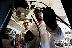 In this photo, a man in a white lab coat and safety glasses attaches a clip to a glass device while another young man looks on in the background.
