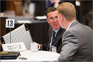 In this photo, two men in suits are at a table. One man holds a sample of his product, which is about the size of a checkerboard and contains membranes to facilitate heating and cooling.