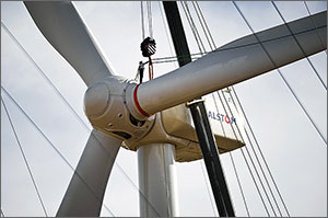 In this photo, a worker is shown standing on top of the rotor of a wind turbine while a crane lifts a blade for the turbine into place.