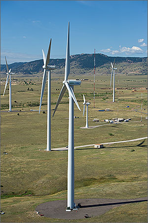 A wide aerial view of a wind turbine testing site and the landscape surrounding it. One large wind turbine is in the foreground with four other wind turbines behind it, as well as several buildings and met towers. Mountains are in the background.