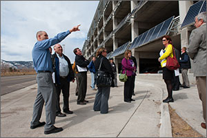 Photo of a man speaking to a group of people standing outside of a parking garage.  Photovoltaic panels are seen on the side of the garage in the background. 