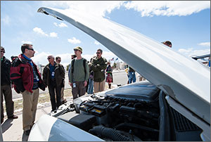 A man stands in front of the open hood of the vehicle.  A group is gathered around him looking at the components of the vehicle under the hood.