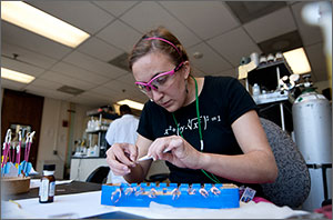 A female works at a laboratory attaching electrodes to a device. In front of her, a blue tray holds more devices waiting for modification.