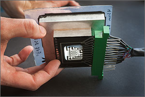 In this photo, a hand holds a test assembly with the glass test card on bottom side, the barrier film on the top, and the metal donut-shaped spacer in the middle. The assembly is the size and shape of a double deck of cards. A cable connects to the test card on one side.
