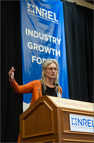 In this photo, a woman in an orange cardigan sweater gestures with her right arm as she stands behind a podium. In the background is a blue banner that reads 