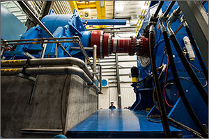 Two large blue devices that make up the dynamometer test system are in the foreground. A large red gearing system, a high speed shaft, connects the two. Behind the device a man in a hard hat is looking at the system.