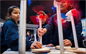 Three girls learn about wind energy by assembling and operating wind turbine models. The blades spin on five wind turbine models in front of the girls.