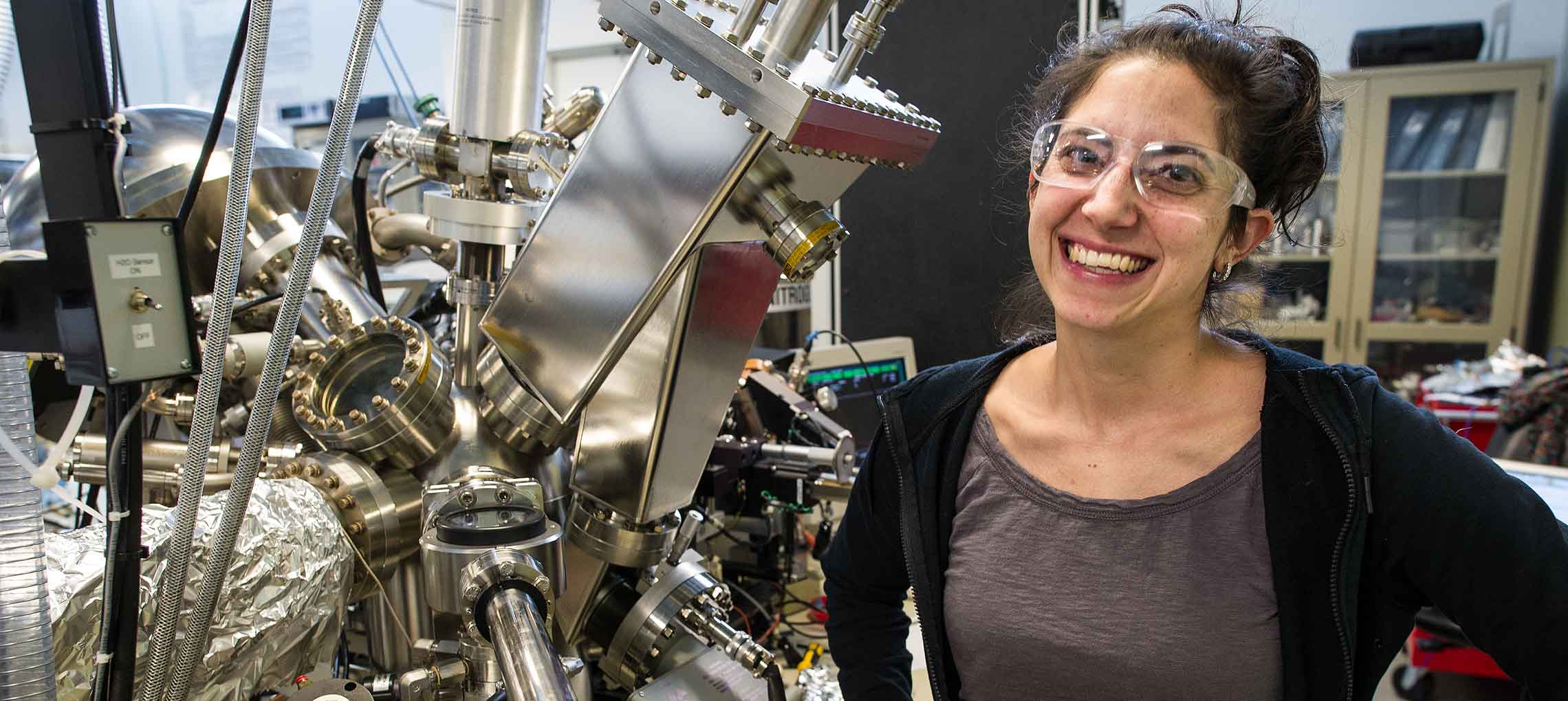 Picture of a smiling woman standing in a room filled with equipment that has many pipes.