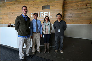 Four smiling people—three men and a woman—stand in front of a wooden wall in an office.