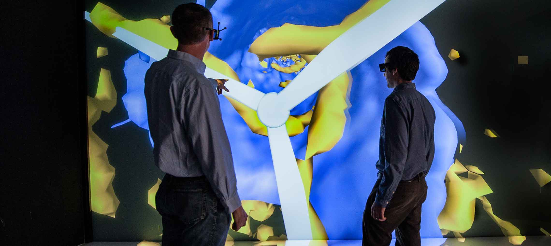 In this photo, a scientist wearing special glasses points to an image on a large visualization screen while another scientist wearing similar glasses looks on.