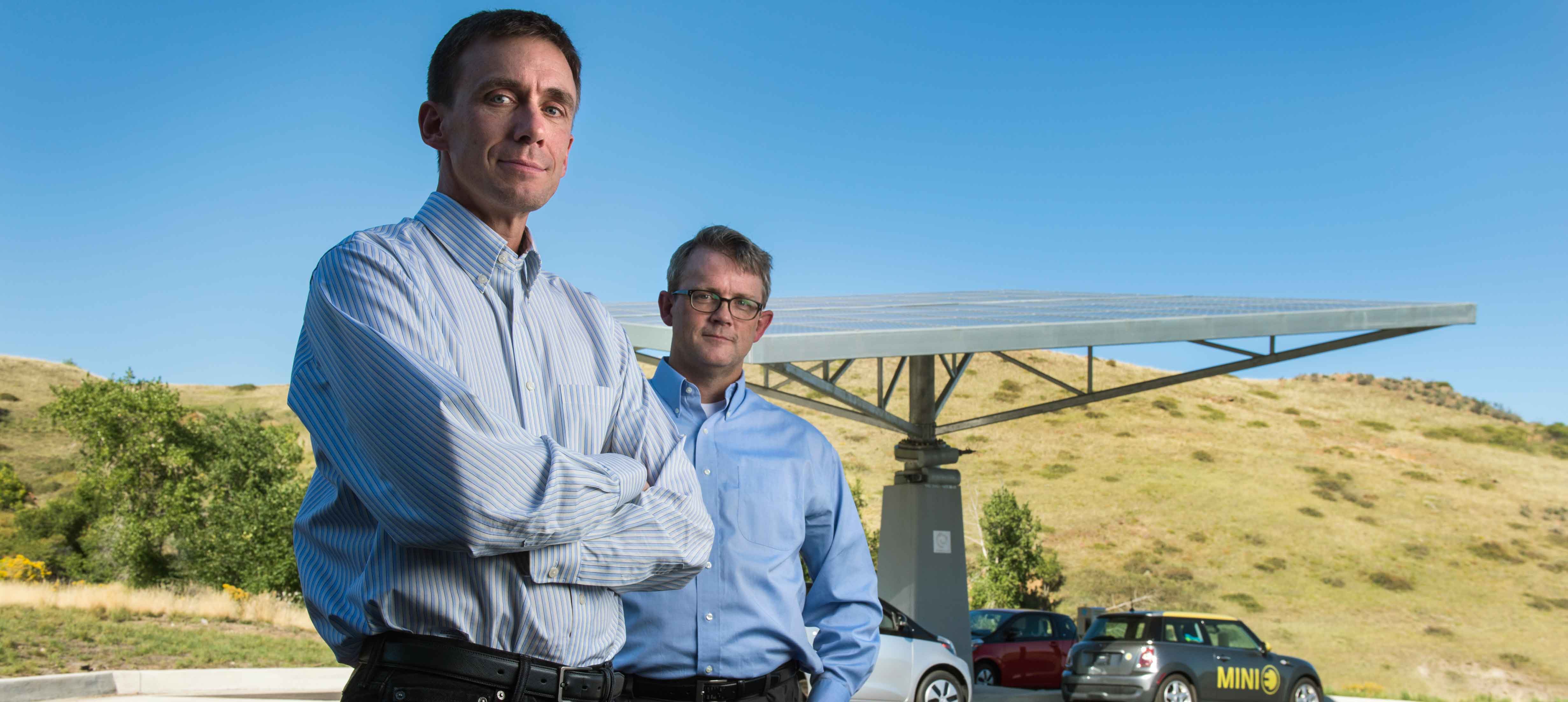 Photo of two men standing in front of a large solar panel and an electric vehicle. 