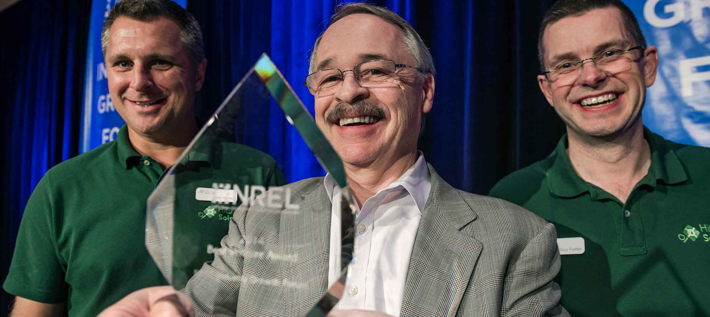 Close-up of three men smiling, the middle one holding a see-through trophy.