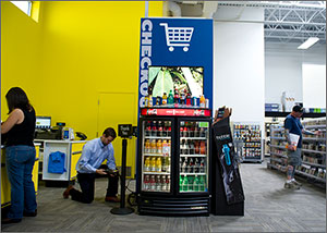 Photo of a man on one knee looking at a portable monitor that is plugged into a chilled display of soft drinks and juices. In the background, a customer at Best Buy peruses merchandise.