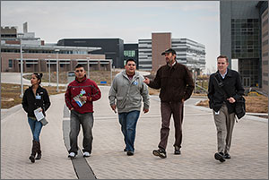 Photo shows a woman and four men walking through NREL's campus on a chilly day, with laboratory buildings in the background.