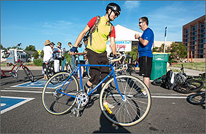 Photo of a man in a bicycle helmet walking his blue road bike past a tent that is serving as a breakfast location for bicyclists at Bike to Work Day at NREL.