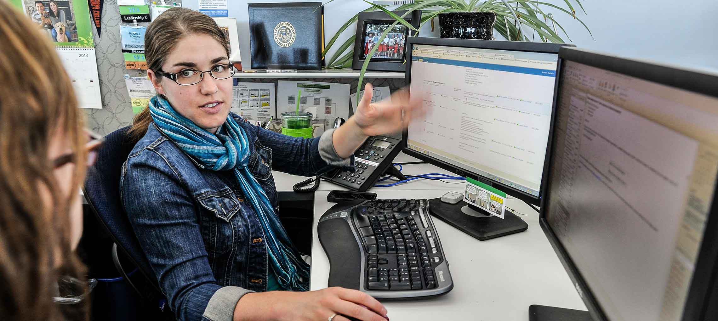 Photo of a woman sitting at a desk with a keyboard, computer screen, and telephone, using her hands to make a point to a co-worker.