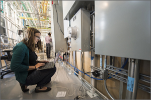 Photo of a woman in a laboratory kneeling in front of a power meter with a computer on her lap.