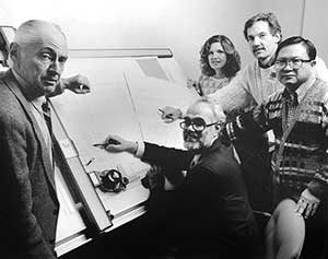 Black and white photo of a group gathered around a drawing board.