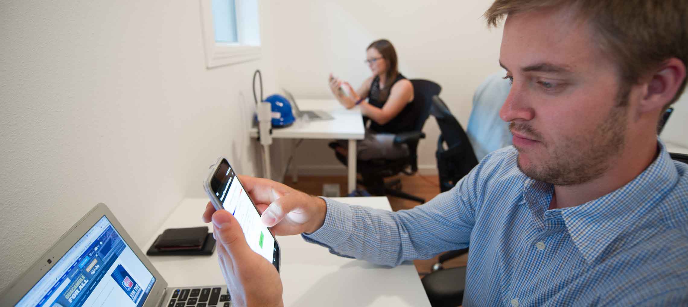 Photo shows two people sitting in a white room, holding smartphones in front of laptops.