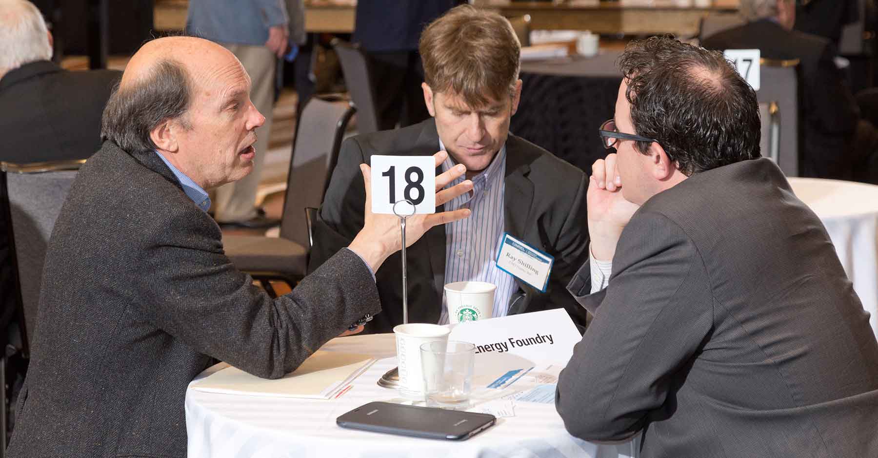 Photo of three men holding a conversation as they sit at a small table.