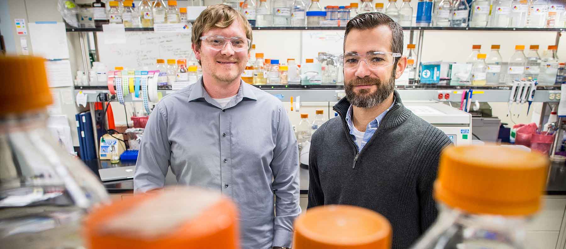 Two men stand in a laboratory filled with glass bottles.