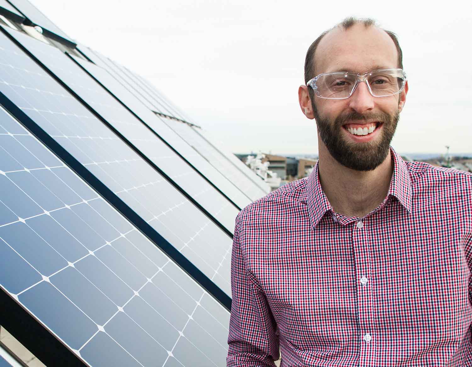 A man wearing safety glasses stands next to large solar panels.