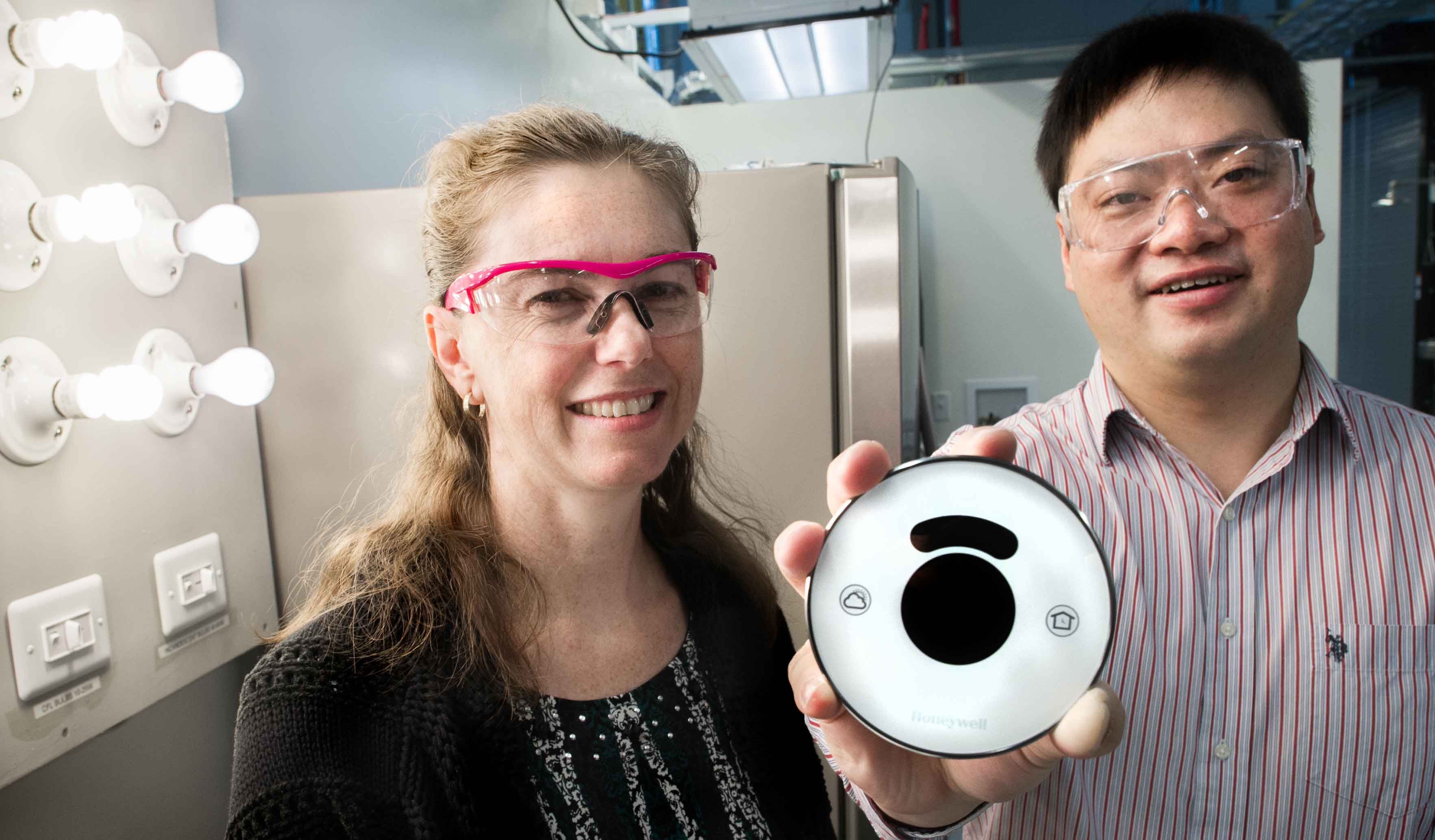 A man and a woman stand inside a laboratory. The man is holding a smart thermostat.