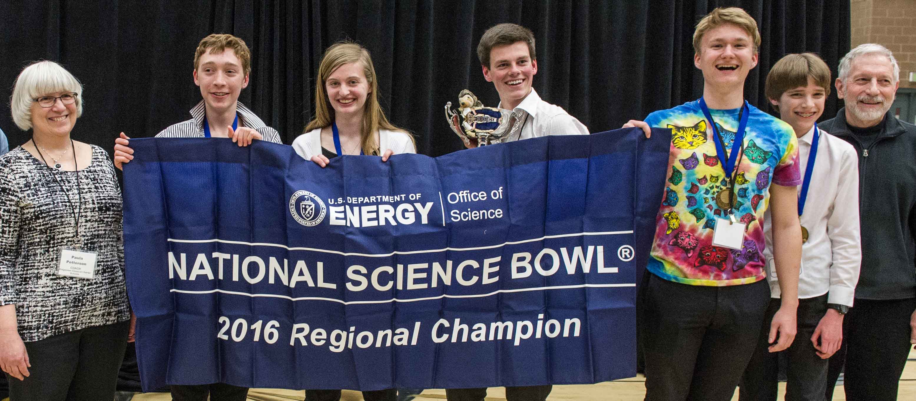 The five student team members from the winning team hold a trophy and banner honoring their victory. Their coaches stand on on each side of the group.